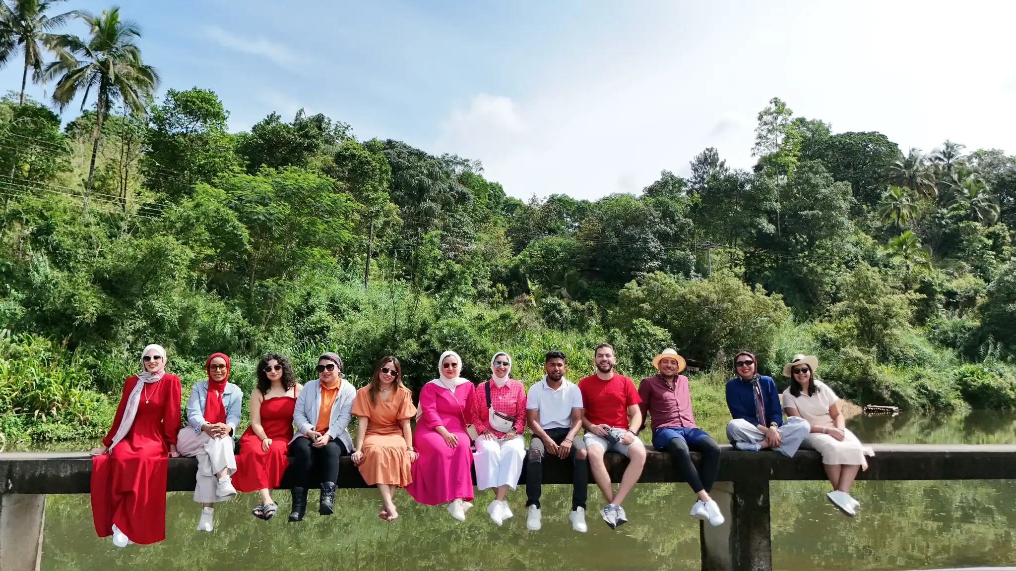 A group sits on a concrete ledge over calm water, surrounded by lush greenery and tropical trees, dressed in colorful casual outfits