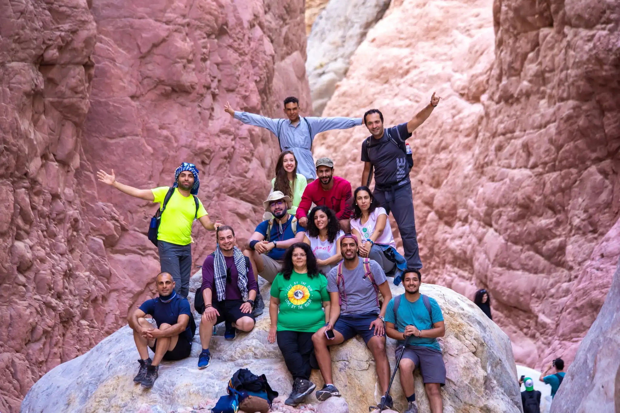 A group of hikers posing on rocks in a narrow canyon with reddish cliffs, wearing casual hiking gear. Some stand with arms raised, while others sit, with more hikers in the background